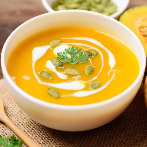 Eine cremige Keto Kürbis Suppe mit Butternut in einer weißen Schüssel, garniert mit Kürbiskernen, frischem Koriander und einem Schuss Kokosmilch, serviert auf einem Holztisch.