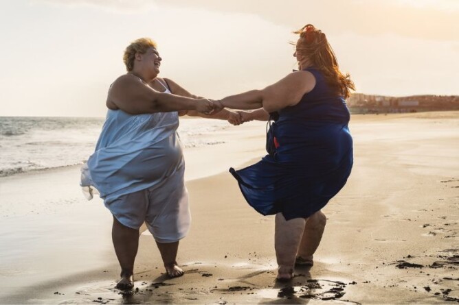 Zwei Frauen tanzen lachend am Strand und symbolisieren Lebensfreude und Selbsthilfe trotz Lipödem.